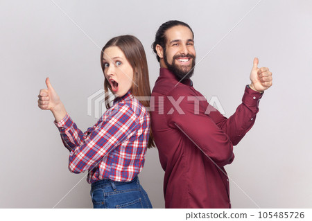 Portrait of young adult woman and man standing back to back together showing thumbs up, like gesture, positive feedback, looking at camera. Indoor studio shot isolated on gray background. Portrait of young adult woman and man standing back to back together showing thumbs up, like gesture, positive feedback, looking at camera. Indoor studio shot isolated on gray background. 105485726