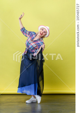 Full length portrait of happy smiling senior woman wearing hat and eyeglasses posing and looking at camera with happiness, dancing. Indoor studio shot isolated on yellow background Full length portrait of happy smiling senior woman wearing hat and eyeglasses posing and looking at camera with happiness, dancing. Indoor studio shot isolated on yellow background 105485727