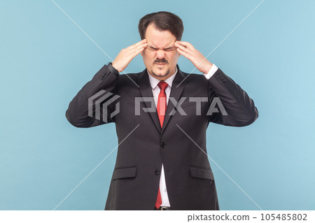 Sick ill man with mustache standing feels unwell, suffering headache, frowning face, massaging temples, wearing black suit with red tie. Indoor studio shot isolated on light blue background. 105485802