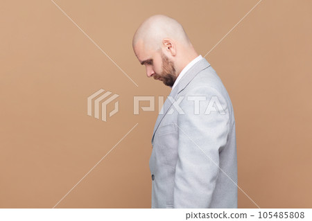 Side view portrait of sad upset bald bearded man standing with head down, being offended, expressing sorrow and sadness, wearing gray jacket. Indoor studio shot isolated on brown background. Side view portrait of sad upset bald bearded man standing with head down, being offended, expressing sorrow and sadness, wearing gray jacket. Indoor studio shot isolated on brown background. 105485808