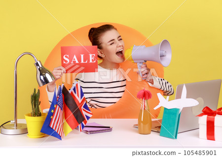 Woman employee of travel agency sitting in rubber ring on neck, showing sale card and screaming in megaphone, hot tours to different countries. Indoor studio studio shot isolated on yellow background. Woman employee of travel agency sitting in rubber ring on neck, showing sale card and screaming in megaphone, hot tours to different countries. Indoor studio studio shot isolated on yellow background. 105485901
