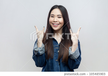 Portrait of smiling happy positive woman in blue denim jacket standing with rock and roll gesture, being in good mood, listens heavy metal, smiling. Indoor studio shot isolated on gray background. 105485903