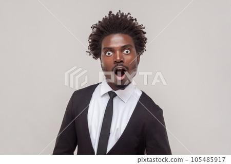 Shocked man with Afro hairstyle stares bugged eyes, feels anxious, looks in disbelief, hears something impressive, wearing white shirt and tuxedo. Indoor studio shot isolated on gray background. Shocked man with Afro hairstyle stares bugged eyes, feels anxious, looks in disbelief, hears something impressive, wearing white shirt and tuxedo. Indoor studio shot isolated on gray background. 105485917
