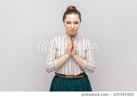 Portrait of cunning young adult woman wearing striped shirt and green skirt keeps hands together, having slander plan, pondering. Indoor studio shot isolated on gray background. 105485920