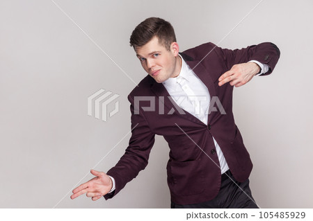 Portrait of attractive positive handsome young man making finger guns and pointing at copy space for advertisement, wearing violet suit and white shirt. Indoor studio shot isolated on grey background. Portrait of attractive positive handsome young man making finger guns and pointing at copy space for advertisement, wearing violet suit and white shirt. Indoor studio shot isolated on grey background. 105485929