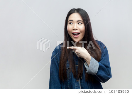 Shocked woman in blue denim jacket standing pointing aside, showing space for your advertisement or promo, promotes big sale has special offer. Indoor studio shot isolated on gray background. Shocked woman in blue denim jacket standing pointing aside, showing space for your advertisement or promo, promotes big sale has special offer. Indoor studio shot isolated on gray background. 105485930