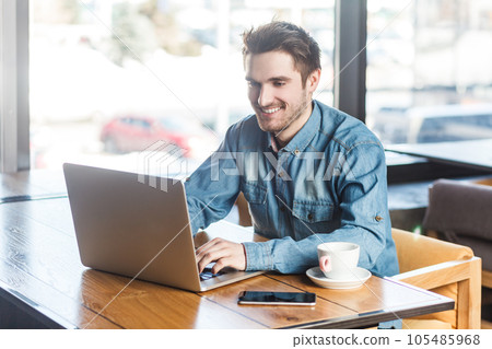 Portrait of smiling happy satisfied young man freelancer in blue jeans shirt working on laptop, typing on keyboard, having video call. Indoor shot near big window, cafe background. Portrait of smiling happy satisfied young man freelancer in blue jeans shirt working on laptop, typing on keyboard, having video call. Indoor shot near big window, cafe background. 105485968