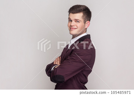 Side view portrait of smiling optimistic young man standing looking ahead, expressing happiness, keeps arms crossed, wearing violet suit and white shirt. Indoor studio shot isolated on grey background Side view portrait of smiling optimistic young man standing looking ahead, expressing happiness, keeps arms crossed, wearing violet suit and white shirt. Indoor studio shot isolated on grey background 105485971