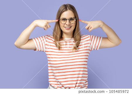 Portrait of delighted young adult blond woman wearing striped T-shirt standing and pointing at her spectacles, looking at camera with optimistic look. Indoor studio shot isolated on purple background. Portrait of delighted young adult blond woman wearing striped T-shirt standing and pointing at her spectacles, looking at camera with optimistic look. Indoor studio shot isolated on purple background. 105485972