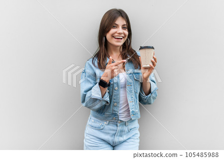 Portrait of joyful cheerful woman wearing denim jacket standing pointing at takeaway coffee, looking at camera with smile, enjoying hot beverage. Indoor studio shot isolated on gray background. Portrait of joyful cheerful woman wearing denim jacket standing pointing at takeaway coffee, looking at camera with smile, enjoying hot beverage. Indoor studio shot isolated on gray background. 105485988