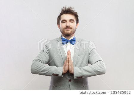 Portrait of calm relaxed bearded man standing with hands together, showing praying gesture, practicing yoga, wearing grey suit and blue bow tie. Indoor studio shot isolated on gray background. 105485992