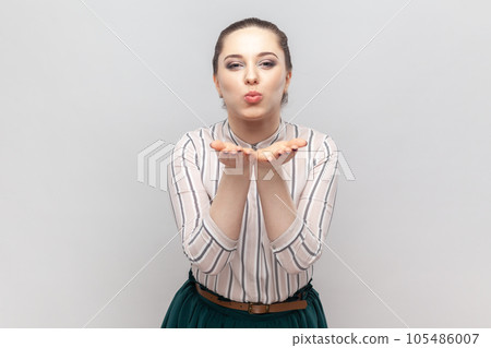 Portrait of romantic flirting woman wearing striped shirt standing sending air kisses over palms, looking at camera, keeps lips pout. Indoor studio shot isolated on gray background. 105486007
