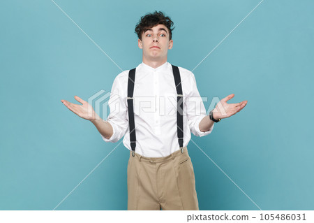 Portrait of puzzled confused man wearing white shirt and suspender, shrugging shoulders, looking away, raised his arms, has uncertain expression. Indoor studio shot isolated on blue background. 105486031