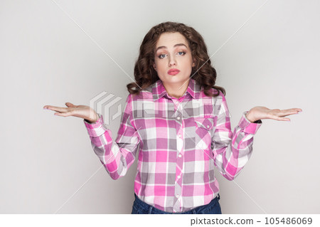 I do not know. Uncertain woman with curly hair shrugging shoulders, keeps palms sideways, has questioned perplexed face expression, no idea what to do. Indoor studio shot isolated on gray background. I do not know. Uncertain woman with curly hair shrugging shoulders, keeps palms sideways, has questioned perplexed face expression, no idea what to do. Indoor studio shot isolated on gray background. 105486069