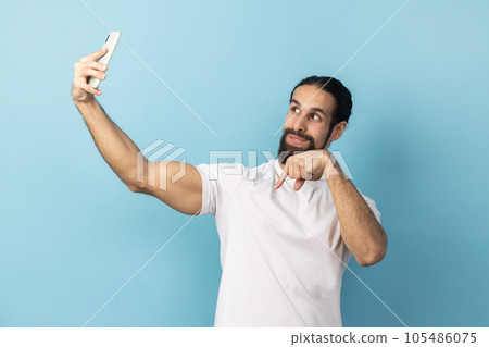 Portrait of optimistic handsome man with beard wearing white T-shirt broadcasting livestream or having video call, pointing finger down, subscribe. Indoor studio shot isolated on blue background. Portrait of optimistic handsome man with beard wearing white T-shirt broadcasting livestream or having video call, pointing finger down, subscribe. Indoor studio shot isolated on blue background. 105486075