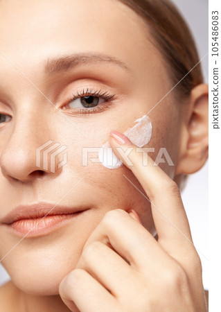Closeup portrait of winsome beautiful woman touching her cheek applying cream grooming her facial skin, cosmetology concept. Indoor studio shot isolated over gray background. 105486083