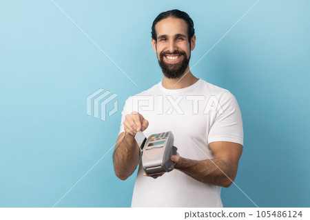 Portrait of man with beard wearing white T-shirt using pos terminal for contactless payment and credit card, paying for shopping, expressing happiness. Indoor studio shot isolated on blue background. 105486124