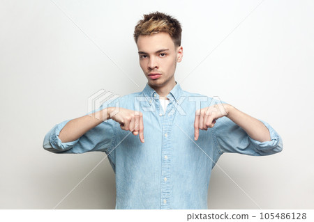 Here and right now. Portrait of strict bossy serious man wearing denim shirt standing looking at camera, pointing down with index fingers. Indoor studio shot isolated on gray background. 105486128