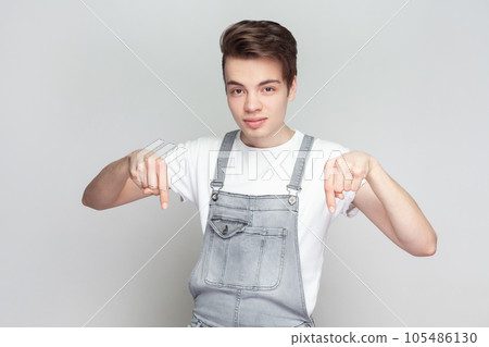Just look down. Portrait of self confident serious young brunette man standing points down, advertises something, wearing denim overalls. Indoor studio shot isolated on gray background. 105486130