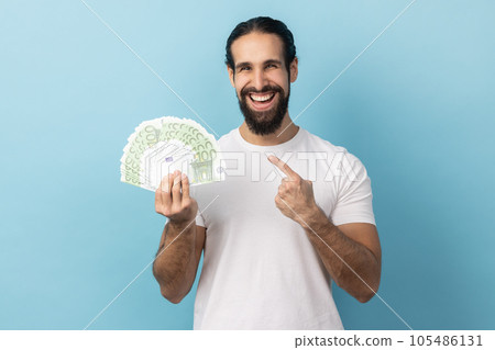 Portrait of satisfied man with beard wearing white T-shirt pointing at fan of euro banknotes, looking at camera with happy expression. Indoor studio shot isolated on blue background. Portrait of satisfied man with beard wearing white T-shirt pointing at fan of euro banknotes, looking at camera with happy expression. Indoor studio shot isolated on blue background. 105486131