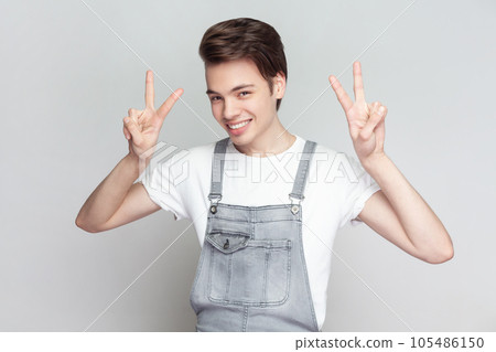 Portrait of young brunette man standing makes peace gesture, shows v sign, looking at camera with toothy smile, wearing denim overalls. Indoor studio shot isolated on gray background. 105486150