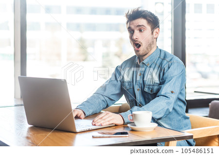 Portrait of amazed astonished surprised man freelancer in blue jeans shirt working on laptop, having video conference, looking at display with big eyes. Indoor shot near big window, cafe background. 105486151