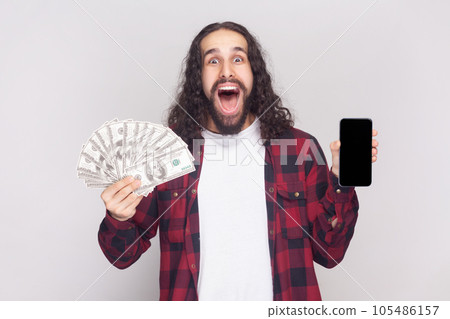 Portrait of amazed excited bearded man with long curly hair in checkered red shirt holding dollar banknotes and cell phone with mockup blank display. Indoor studio shot isolated on gray background. 105486157