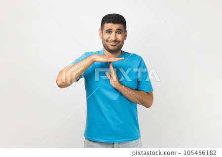 Portrait of attractive young adult unshaven man wearing blue T- shirt standing showing time out gesture with hands, looking at camera. Indoor studio shot isolated on gray background. 105486182