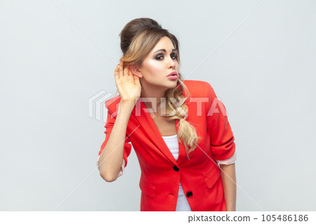 Portrait of curious blonde woman standing holding palm near ear, having problems with hearing, listening to gossip, wearing red jacket. Indoor studio shot isolated on gray background. Portrait of curious blonde woman standing holding palm near ear, having problems with hearing, listening to gossip, wearing red jacket. Indoor studio shot isolated on gray background. 105486186