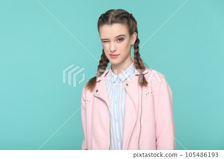 Portrait of playful cheerful teenager girl with braids wearing pink jacket standing looking at camera, winking, flirting with boyfriend. Indoor studio shot isolated on green background. Portrait of playful cheerful teenager girl with braids wearing pink jacket standing looking at camera, winking, flirting with boyfriend. Indoor studio shot isolated on green background. 105486193