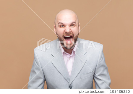 Portrait of angry or shocked bald bearded man standing, looking at camera and screaming, arguing with friend, wearing gray jacket. Indoor studio shot isolated on brown background. Portrait of angry or shocked bald bearded man standing, looking at camera and screaming, arguing with friend, wearing gray jacket. Indoor studio shot isolated on brown background. 105486220