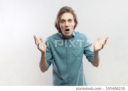Portrait of shocked excited worried young man raised his arms, looking at camera, sees something unbelievable, saying wow, wearing blue shirt. Indoor studio shot isolated on gray background. Portrait of shocked excited worried young man raised his arms, looking at camera, sees something unbelievable, saying wow, wearing blue shirt. Indoor studio shot isolated on gray background. 105486238