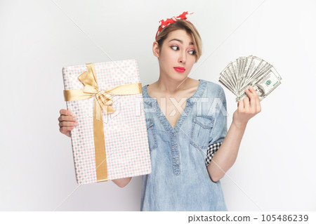 Portrait of confused puzzled uncertain blonde woman wearing blue denim shirt and red headband standing holding present box and dollar banknotes. Indoor studio shot isolated on gray background. 105486239