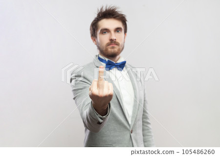 Portrait of rude impolite handsome bearded man standing showing middle finger, arguing with somebody, wearing grey suit and blue bow tie. Indoor studio shot isolated on gray background. 105486260