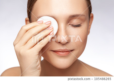 Closeup portrait of beautiful woman with closed eyes standing naked and showing cotton pads near face, wipe-off makeup before shower. Indoor studio shot isolated over gray background. 105486265