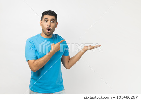Portrait of amazed excited surprised astonished unshaven man wearing blue T- shirt standing presenting advertisement area on his palm, mockup. Indoor studio shot isolated on gray background. 105486267