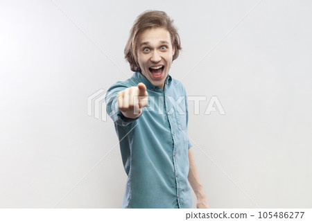 Portrait of surprised astonished shocked young man standing pointing at camera, selecting you, expressing positive emotions, wearing blue shirt. Indoor studio shot isolated on gray background. Portrait of surprised astonished shocked young man standing pointing at camera, selecting you, expressing positive emotions, wearing blue shirt. Indoor studio shot isolated on gray background. 105486277