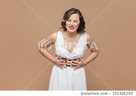 Portrait of unhealthy sick middle aged woman with wavy hair standing, holding her belly with hands, stomach cramps, wearing white dress. Indoor studio shot isolated on light brown background. 105486289