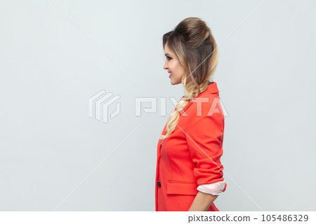 Side view portrait of smiling attractive positive optimistic blonde woman standing looking ahead, expressing positiveness, wearing red jacket. Indoor studio shot isolated on gray background. Side view portrait of smiling attractive positive optimistic blonde woman standing looking ahead, expressing positiveness, wearing red jacket. Indoor studio shot isolated on gray background. 105486329