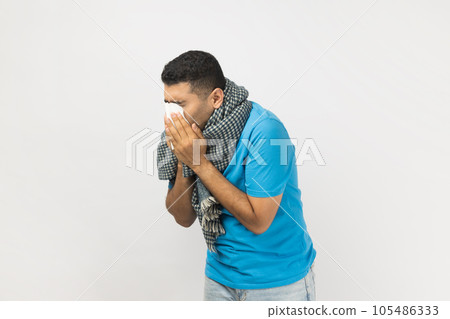 Portrait of ill sick unshaven man wearing blue T- shirt and wrapped in warm scarf standing sneezing, having runny nose, has grippe. Indoor studio shot isolated on gray background. Portrait of ill sick unshaven man wearing blue T- shirt and wrapped in warm scarf standing sneezing, having runny nose, has grippe. Indoor studio shot isolated on gray background. 105486333