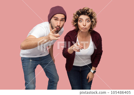 Portrait of astonished couple of friends standing together and pointing at camera, choosing you, looking with big eyes, being amazed. Indoor studio shot isolated on pink background. Portrait of astonished couple of friends standing together and pointing at camera, choosing you, looking with big eyes, being amazed. Indoor studio shot isolated on pink background. 105486405