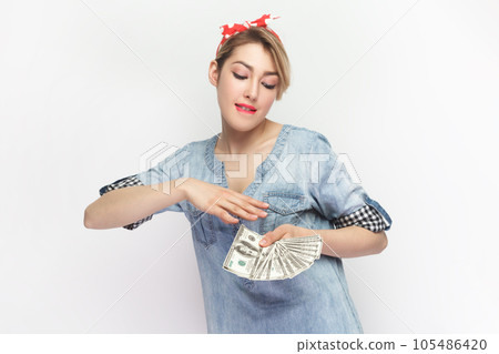 Portrait of rich satisfied blonde attractive woman wearing blue denim shirt and red headband standing throwing money, having big salary. Indoor studio shot isolated on gray background. Portrait of rich satisfied blonde attractive woman wearing blue denim shirt and red headband standing throwing money, having big salary. Indoor studio shot isolated on gray background. 105486420
