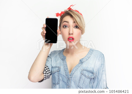 Portrait of shocked blonde young woman wearing blue denim shirt and red headband standing holding smart phone with black blank screen. Indoor studio shot isolated on gray background. 105486421