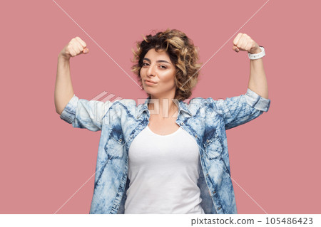 Portrait of self confident strong woman with curly hairstyle wearing blue shirt standing with raised arms, showing her power, looking at camera. Indoor studio shot isolated on pink background. 105486423