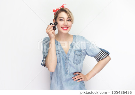 Portrait of smiling cheerful joyful blonde woman wearing blue denim shirt and red headband standing holding spinner, keeps hand on hips. Indoor studio shot isolated on gray background. 105486456