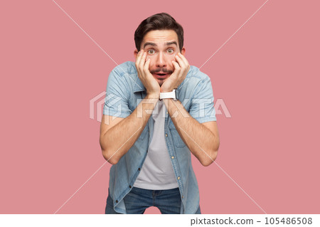 Portrait of shocked astonished bearded man in blue casual style shirt standing with hands on cheeks, looking at camera with big eyes. Indoor studio shot isolated on pink background. 105486508