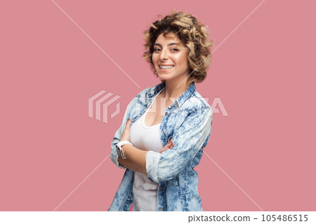 Portrait of confident happy smiling woman with curly hairstyle wearing blue shirt standing with crossed arms, looking at camera with positive expression. Indoor studio shot isolated on pink background Portrait of confident happy smiling woman with curly hairstyle wearing blue shirt standing with crossed arms, looking at camera with positive expression. Indoor studio shot isolated on pink background 105486515