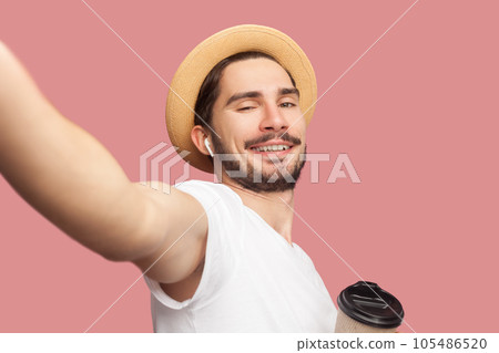 Portrait of handsome cheerful joyful bearded man in white T-shirt and hat standing drinking coffee, looking smiling at camera, POV. Indoor studio shot isolated on pink background. Portrait of handsome cheerful joyful bearded man in white T-shirt and hat standing drinking coffee, looking smiling at camera, POV. Indoor studio shot isolated on pink background. 105486520