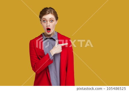 Portrait of amazed astonished woman with red lips standing pointing finger aside at copy space for advertisement, wearing red jacket. Indoor studio shot isolated on yellow background. Portrait of amazed astonished woman with red lips standing pointing finger aside at copy space for advertisement, wearing red jacket. Indoor studio shot isolated on yellow background. 105486525