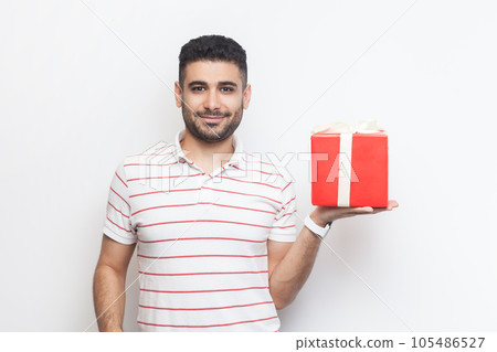 Portrait of delighted cheerful handsome bearded man wearing striped t-shirt standing showing red wrapped present box, gift for birthday. Indoor studio shot isolated on gray background. Portrait of delighted cheerful handsome bearded man wearing striped t-shirt standing showing red wrapped present box, gift for birthday. Indoor studio shot isolated on gray background. 105486527